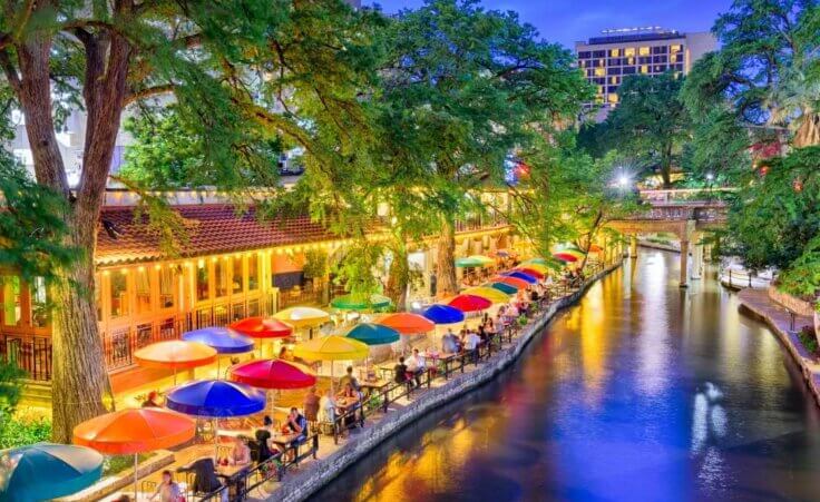 Diners sit at umbrella-covered tables sit alongside the Riverwalk in San Antonio at night