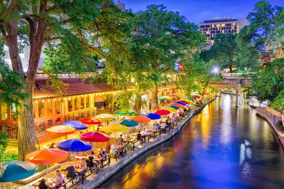 Diners sit at umbrella-covered tables sit alongside the Riverwalk in San Antonio at night
