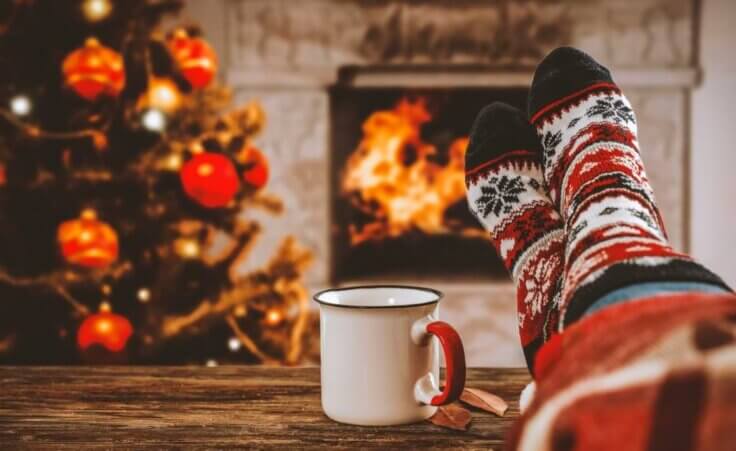 Crossed legs in festive Christmas socks rest atop a wooden table next to a coffee mug and in front of a Christmas tree and fireplace with a roaring fire. © By magdal3na/stock.adobe.com