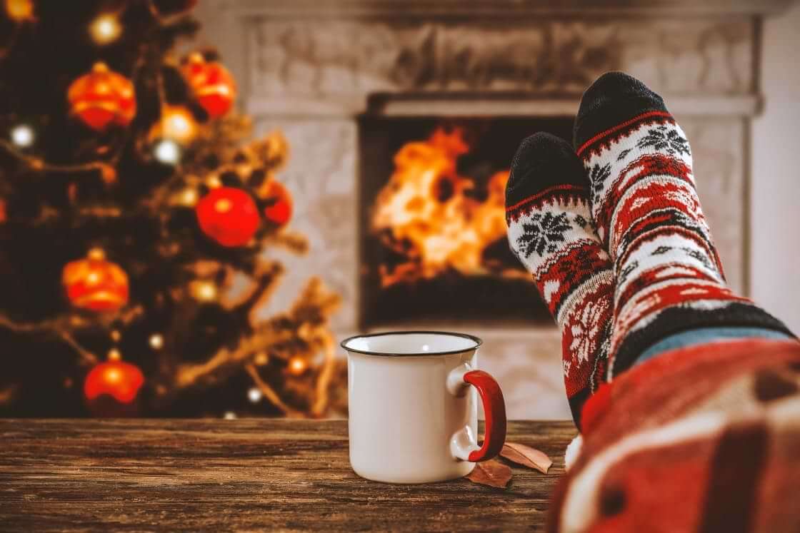 Crossed legs in festive Christmas socks rest atop a wooden table next to a coffee mug and in front of a Christmas tree and fireplace with a roaring fire. © By magdal3na/stock.adobe.com