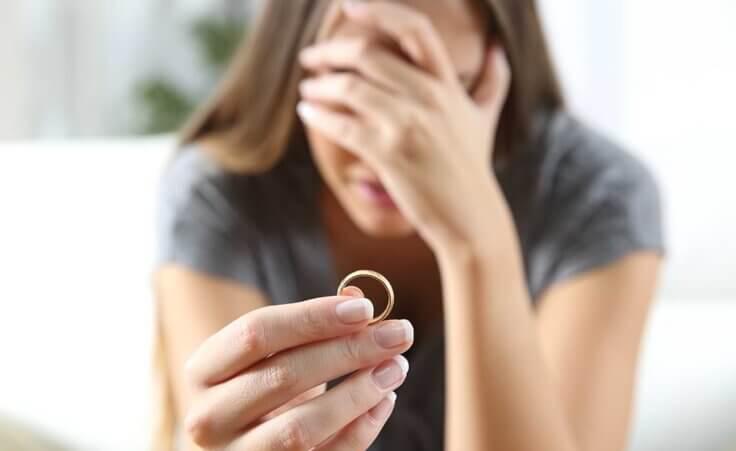 A distraught woman, her hand over her face, holds her wedding ring out in front of her.