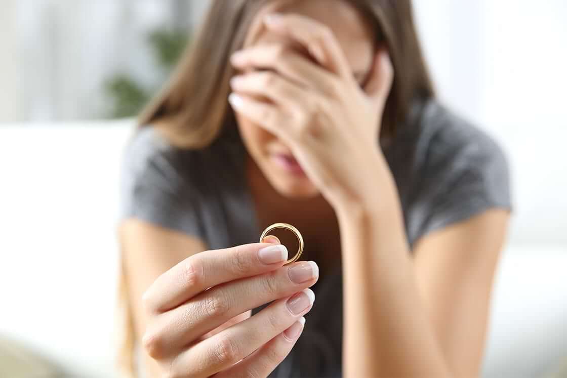 A distraught woman, her hand over her face, holds her wedding ring out in front of her.