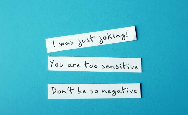 Three phrases denoting gaslighting—"I was just joking! You are too sensitive. Don't be so negative."—are written on three separate strips of white paper set on a blue background.