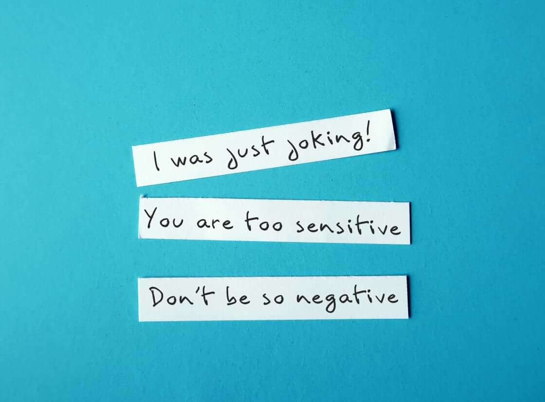 Three phrases denoting gaslighting—"I was just joking! You are too sensitive. Don't be so negative."—are written on three separate strips of white paper set on a blue background.
