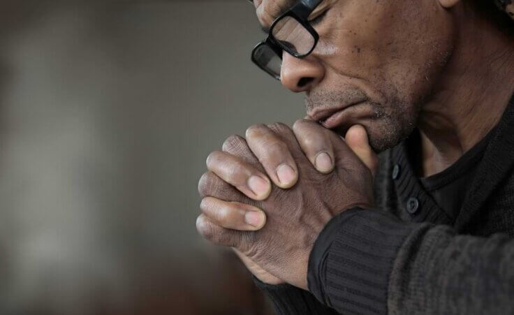 A man prays with his hands clasped below his chin, his eyes shut.