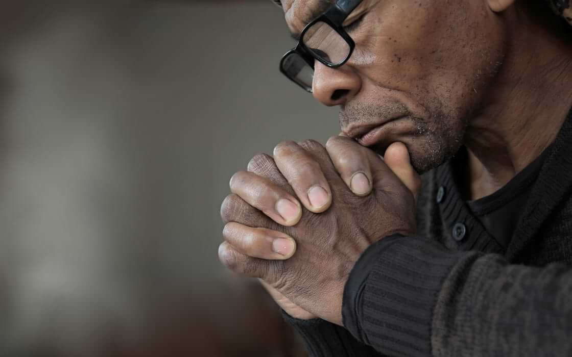 A man prays with his hands clasped below his chin, his eyes shut.