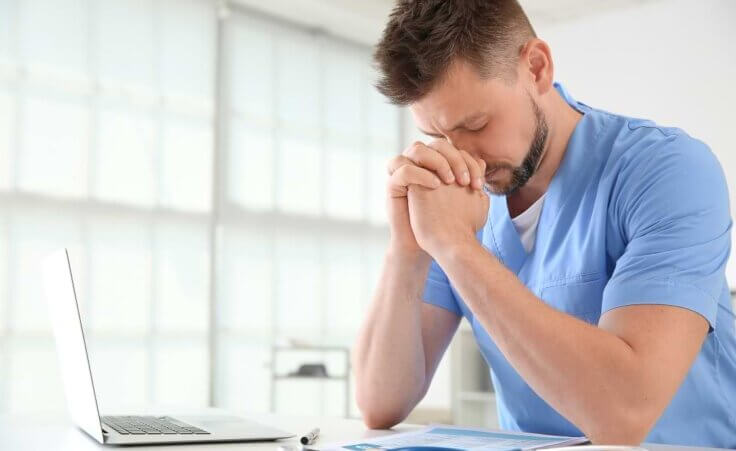 A doctor in blue scrubs sits at a desk in front of an open laptop with his hands clasped in prayer.