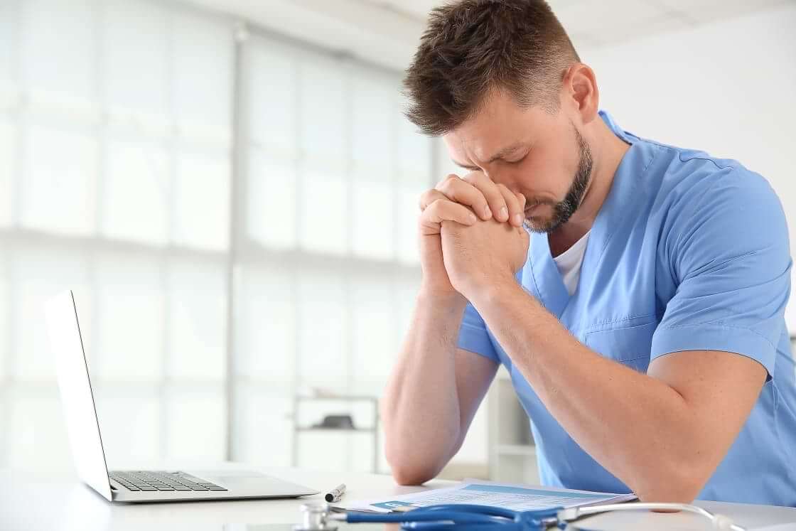 A doctor in blue scrubs sits at a desk in front of an open laptop with his hands clasped in prayer.
