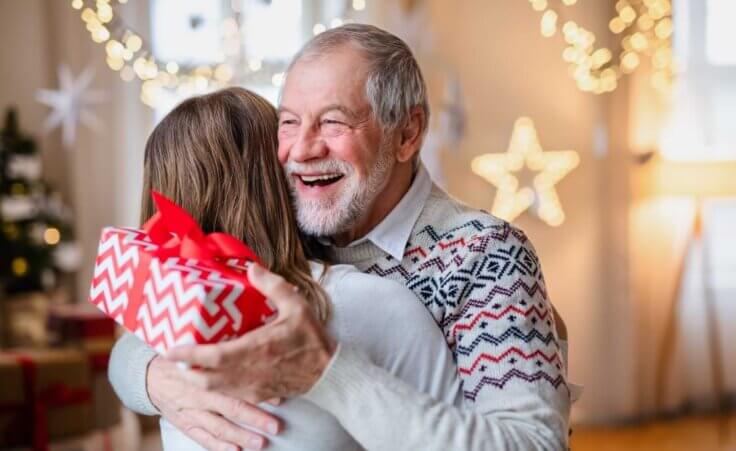 A grandpa holds a Christmas present in one hand as he hugs a woman.