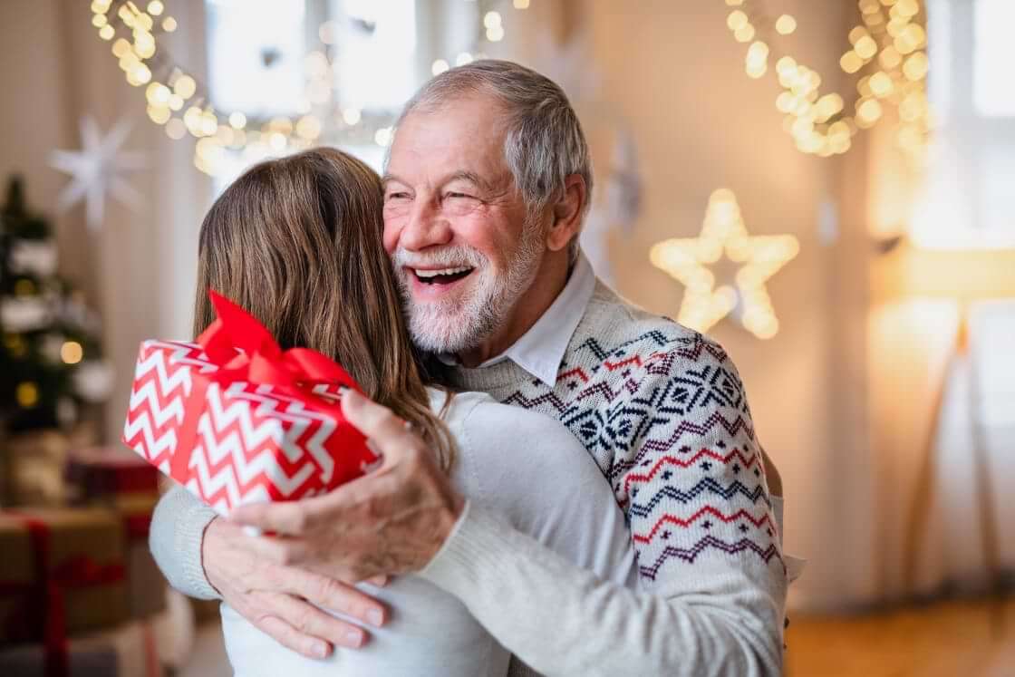 A grandpa holds a Christmas present in one hand as he hugs a woman.