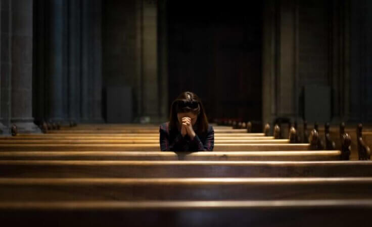 Alone at church, a woman sits in a pew, her head bowed and her hands folded in prayer.
