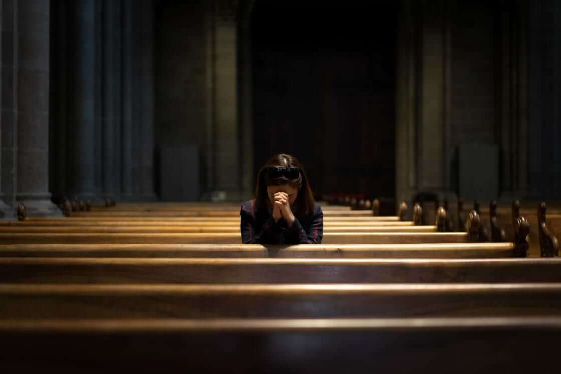 Alone at church, a woman sits in a pew, her head bowed and her hands folded in prayer.