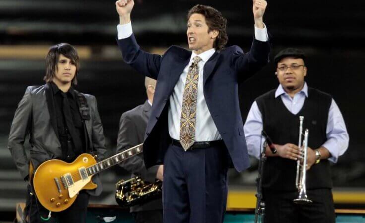 Joel Osteen preaching in front of the church band, with his arms outstretched.