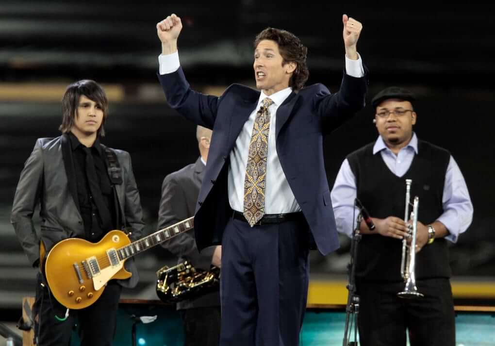 Joel Osteen preaching in front of the church band, with his arms outstretched.