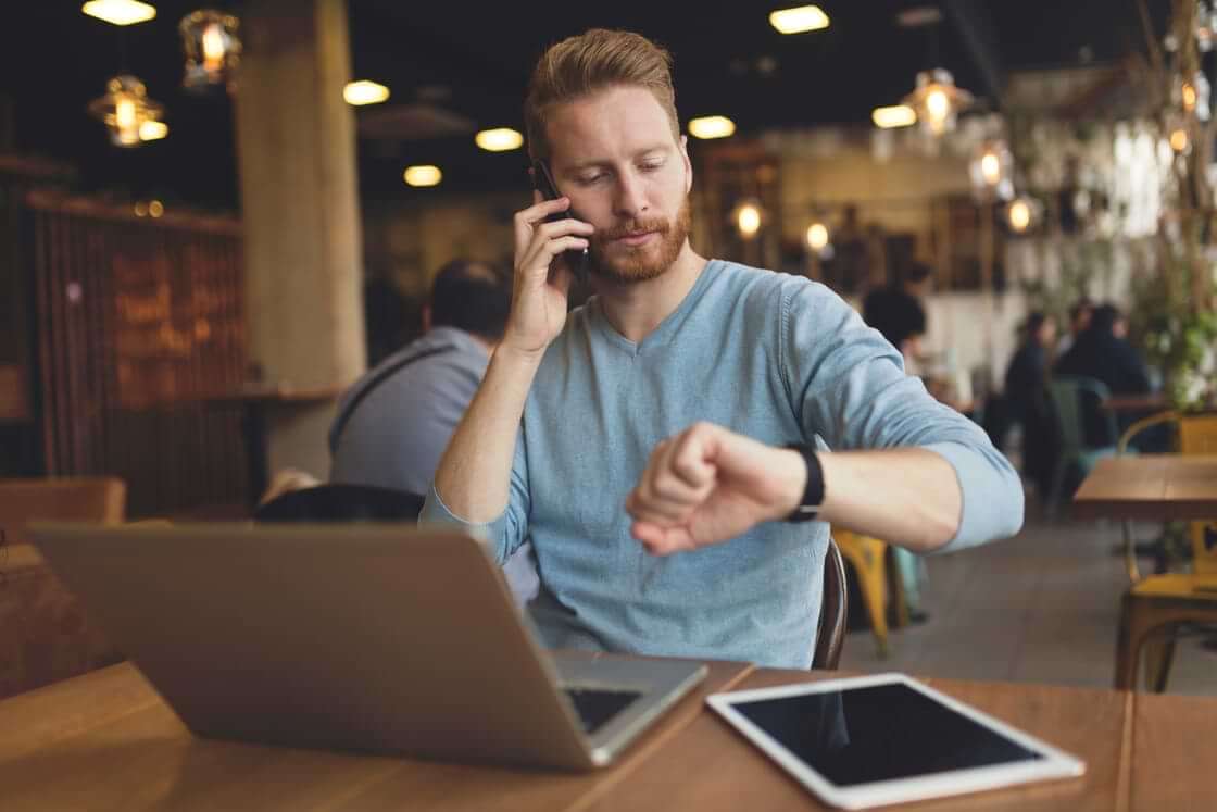 A busy man talks on his phone while looking at his watch while sitting in front of an open laptop at a coffeeshop. © By NDABCREATIVITY/stock.adobe.com