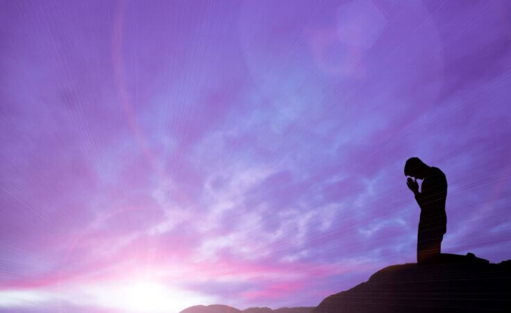 A man prays alone on a mountaintop against a purple-sky sunrise backdrop. © By ctvvelve/stock.adobe.com