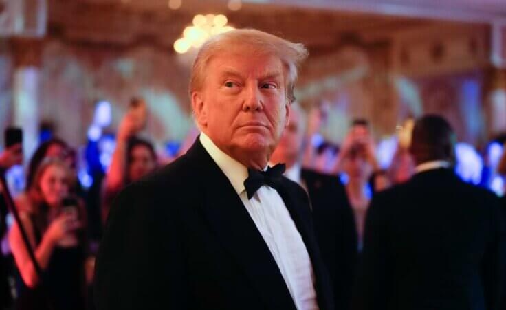 Trump wearing a tuxedo stands in his hotel with people partying behind him.
