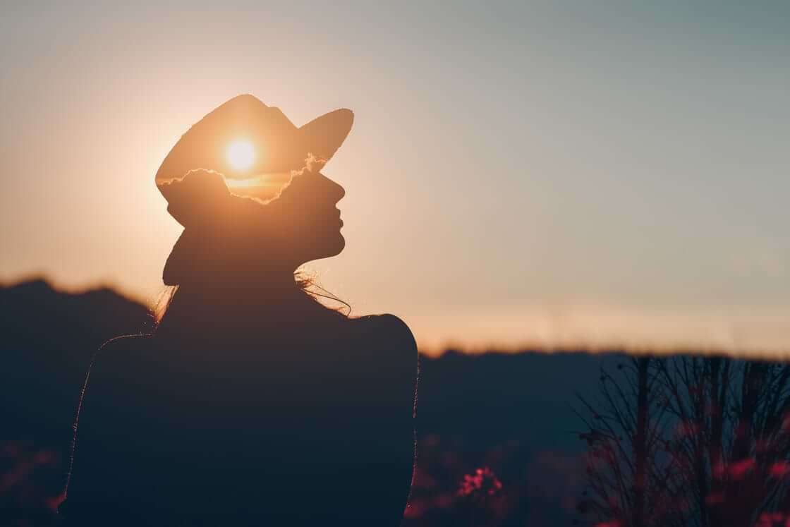 A silhouetted woman in a hat stands outside at sunset, her face looking to the side, her hat artistically filled with the sunset. © By primipil/stock.adobe.com