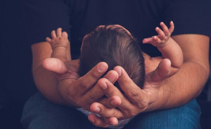 A man holds a baby in his lap, his hands cupped around the child's head, depicting one meaning of being pro-life today. © By Tamara Sales/stock.adobe.com