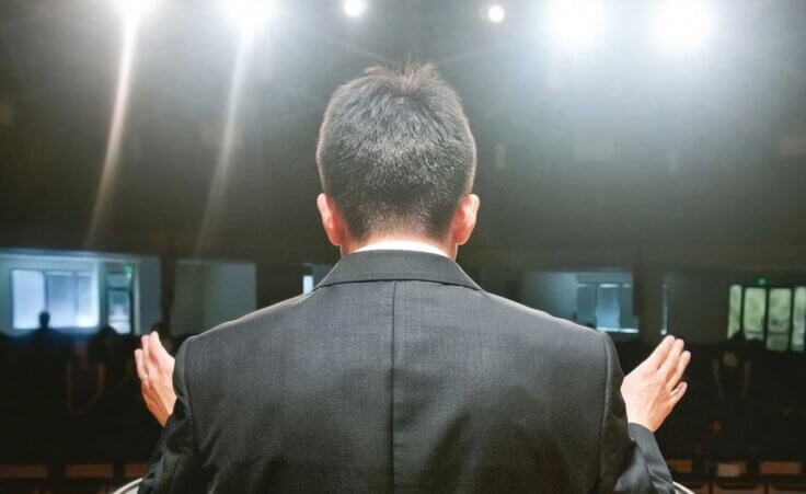 A pastor behind a pulpit raises his hands as spotlights shine down. © By shoenberg3/stock.adobe.com