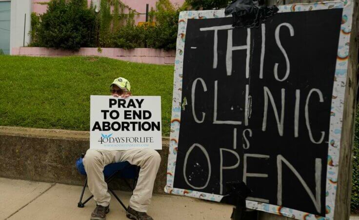 FILE - An anti-abortion supporter sits behind a sign that advises the Jackson Women's Health Organization clinic is still open in Jackson, Miss., Wednesday, July 6, 2022. (AP Photo/Rogelio V. Solis, File)