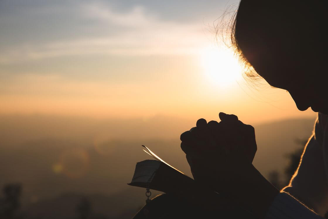 A woman prays with hands clasped over a Bible while outside at sunrise. © By Tinnakorn/stock.adobe.com
