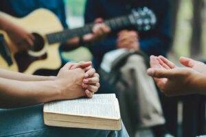 Stock photo: In the foreground, a man clasps his hands in prayer over an open Bible while another pair of hands is open in prayer. A guitarist and another person in prayer with an open Bible are in the background. Similar scenes are occurring at the revival in Asbury and beyond. © By Pcess609/stock.adobe.com