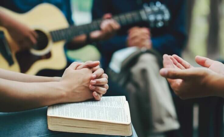 Stock photo: In the foreground, a man clasps his hands in prayer over an open Bible while another pair of hands is open in prayer. A guitarist and another person in prayer with an open Bible are in the background. Similar scenes are occurring at the revival in Asbury and beyond. © By Pcess609/stock.adobe.com