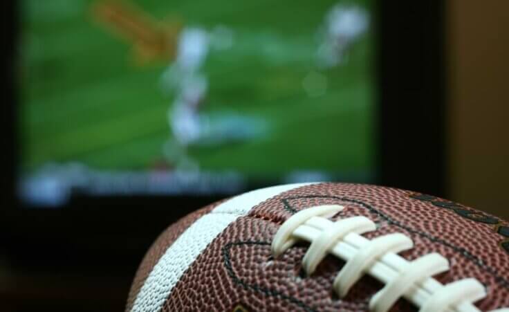 A football sits in front of a TV. The He Gets Us campaign plans to spend nearly $20 million for sixty second of airtime during Super Bowl LVII this Sunday. © By JJAVA/stock.adobe.com