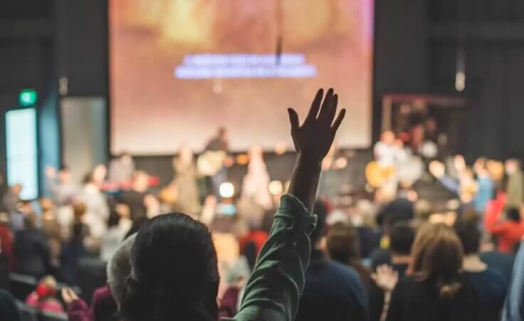 A Christian worshipper raises their hand during a church worship service. © By tutye/stock.adobe.com. Harvard University reports that going to church is associated with a lower risk of a "death of despair."