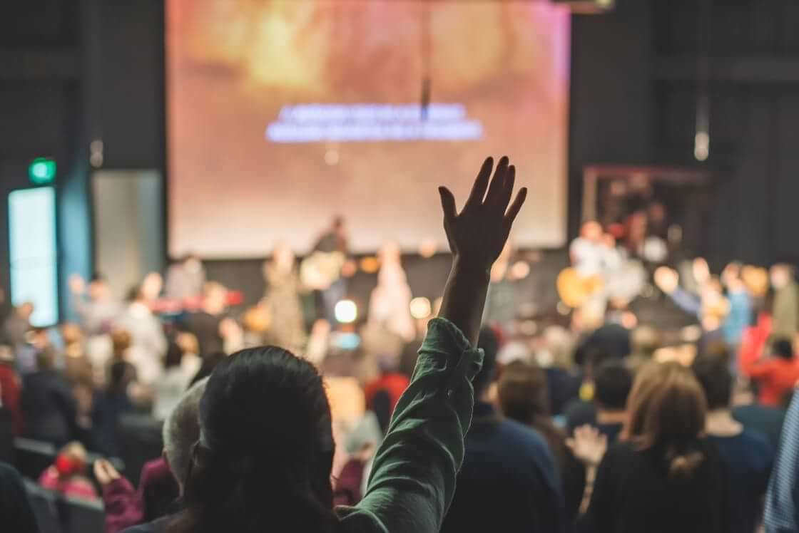 A Christian worshipper raises their hand during a church worship service. © By tutye/stock.adobe.com. Harvard University reports that going to church is associated with a lower risk of a "death of despair."