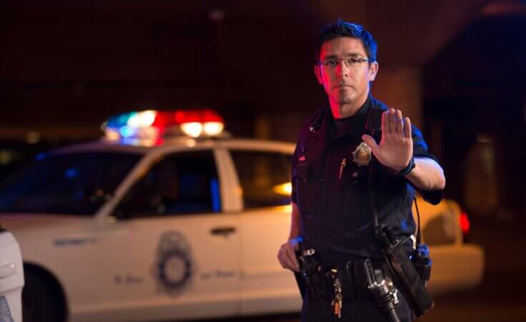 A police officer, standing in front of his car, holds up his left palm in a signal to stop. © By Studio615/stock.adobe.com