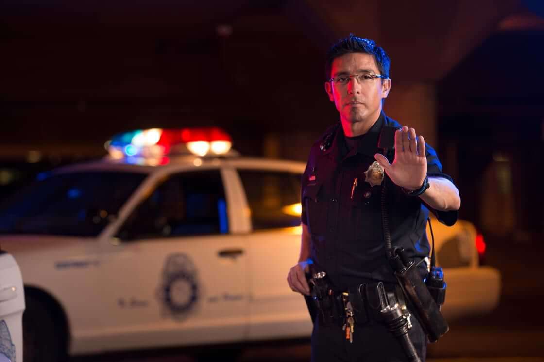 A police officer, standing in front of his car, holds up his left palm in a signal to stop. © By Studio615/stock.adobe.com
