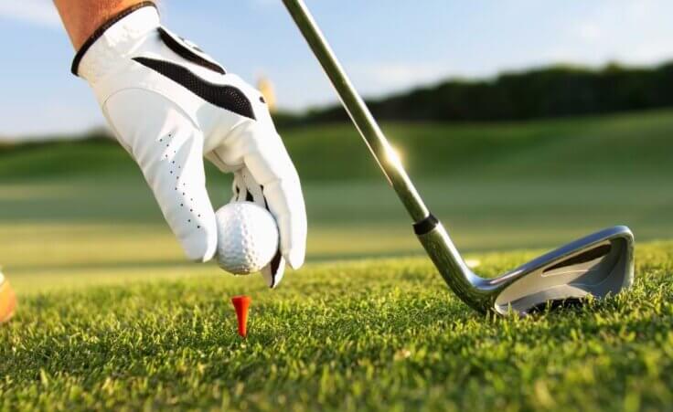 Close-up of a golfer wearing a golf glove placing a golf ball on a tee, with an iron in the background. © By sculpies/stock.adobe.com