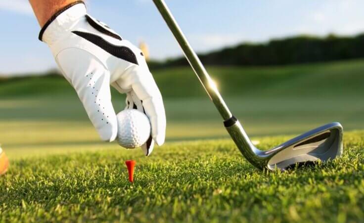 Close-up of a golfer wearing a golf glove placing a golf ball on a tee, with an iron in the background. © By sculpies/stock.adobe.com