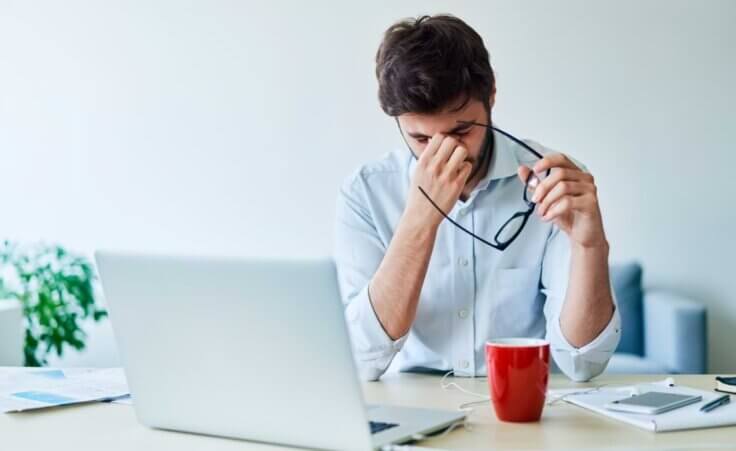 A man in an office, sitting in front of his open laptop, pinches his nose as he removes his glasses in a sign of stress. Many executives, including pastors, experience Toxic Stress Load. © By baranq/stock.adobe.com