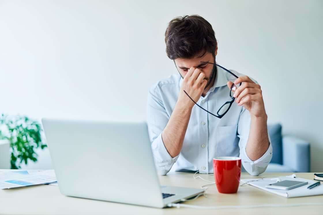 A man in an office, sitting in front of his open laptop, pinches his nose as he removes his glasses in a sign of stress. Many executives, including pastors, experience Toxic Stress Load. © By baranq/stock.adobe.com