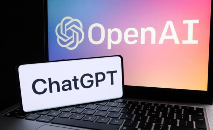 A smartphone displays the word ChatGPT in the foreground while resting on the keyboard of an open laptop displaying the logo of OpenAI. © By Ascannio/stock.adobe.com