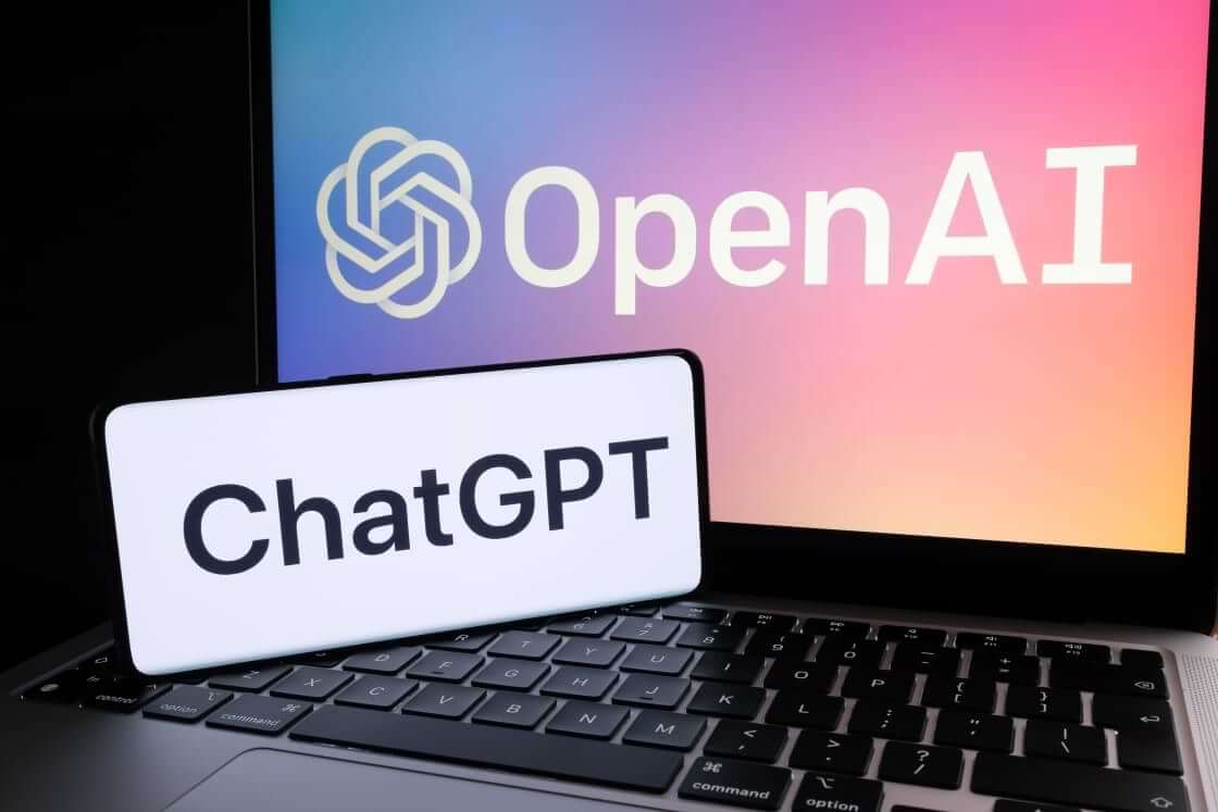 A smartphone displays the word ChatGPT in the foreground while resting on the keyboard of an open laptop displaying the logo of OpenAI. © By Ascannio/stock.adobe.com