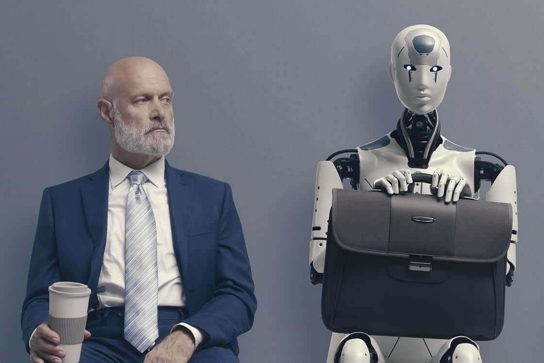 An AI robot sits with a briefcase in its lap next to a man in a suit, both of them waiting for a job interview. Elon Musk and 1000 tech experts are calling for an AI moratorium of 6 months. © By stokkete/stock.adobe.com