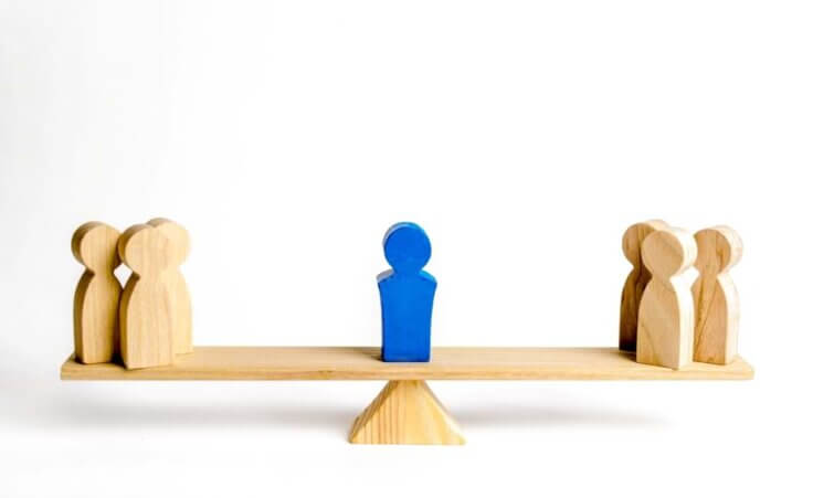 Seven wooden figures are perched on a wooden seesaw, three on the left, three on the right, and one painted blue in the middle symbolizing conflict avoidant pastors and church leaders. © By Andrii Yalanskyi/stock.adobe.com