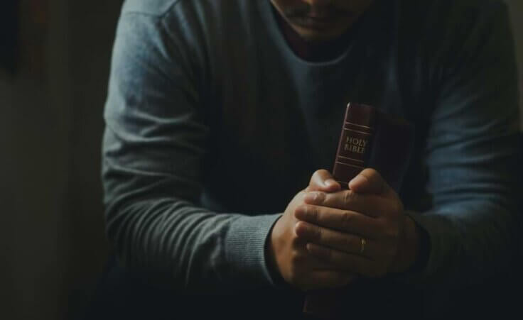 A sitting man holds a Bible between his hands as he rests his arms on his legs. He may be wrestling with faith and doubt. © By Pcess609/stock.adobe.com