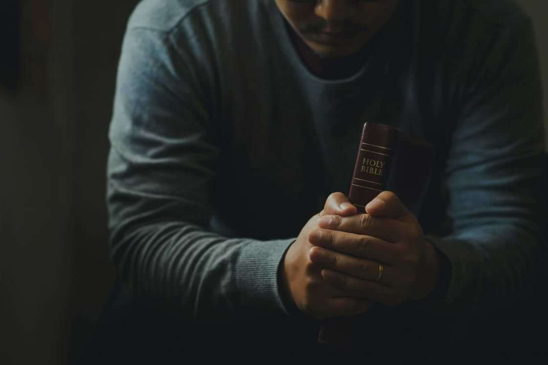 A sitting man holds a Bible between his hands as he rests his arms on his legs. He may be wrestling with faith and doubt. © By Pcess609/stock.adobe.com