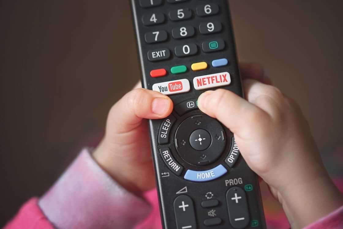 Preschool-age hands hold a remote displaying buttons for YouTube and Netflix. Ridley Jones, a Netflix series for preschoolers, recently featured a nonbinary character coming out and desiring to be called "they." © By Tricky Shark/stock.adobe.com