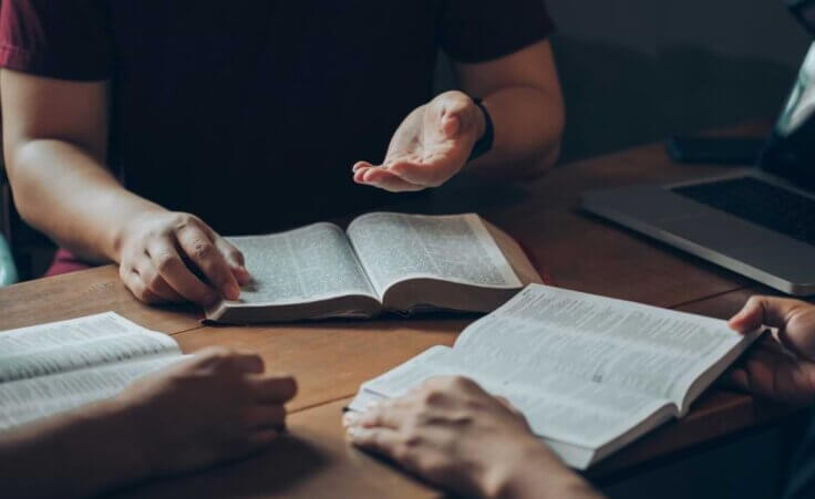 Three people in a small group Bible study are gathered around a table, all with Bibles open. © By Pcess609/stock.adobe.com