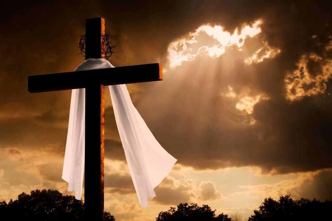 Against a background of the sun breaking through the clouds stand an empty cross adorned with a crown of thorns and draped with white fabric, a reminder of the great cost Jesus paid on Good Friday. © By ricardoreitmeyer/stock.adobe.com