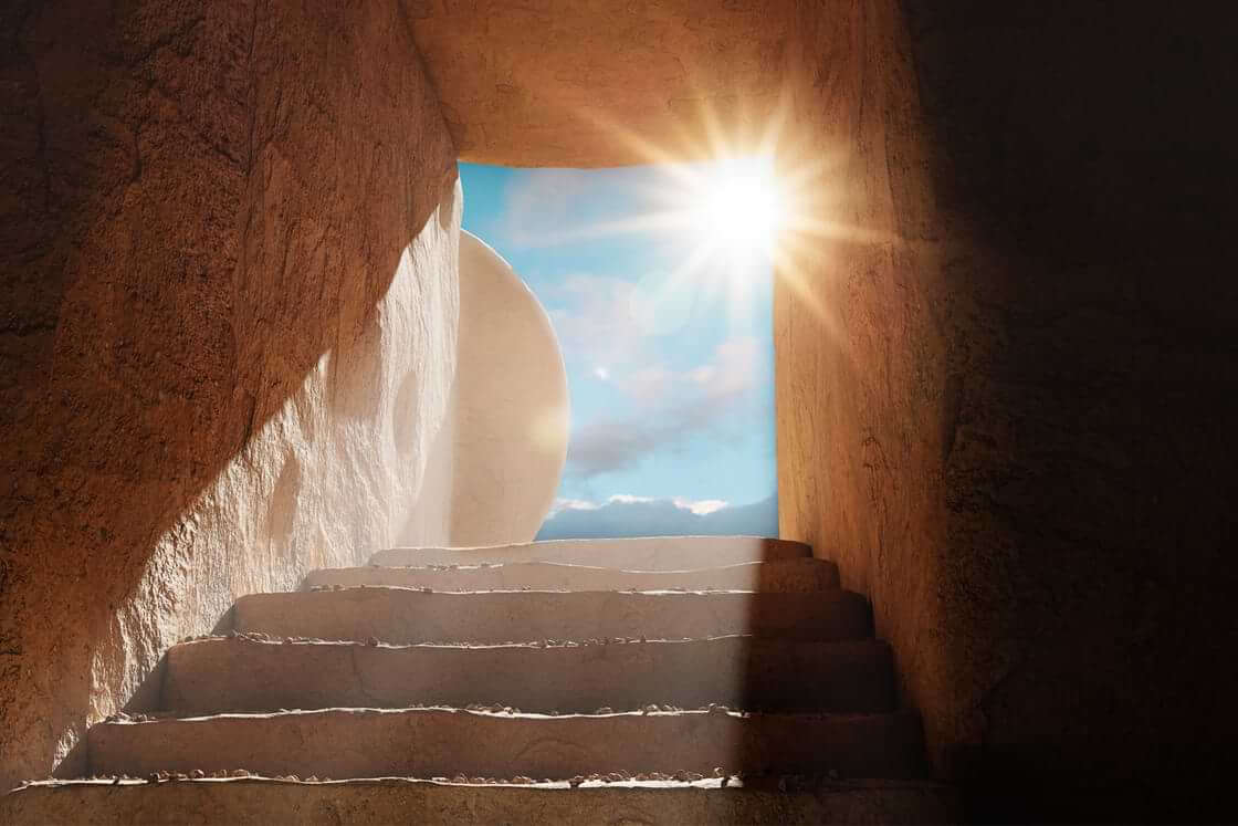 Steps within a dark tomb lead up to an opening where a stone has been rolled away as a bright sun shines through, a scene evoking Easter Sunday morning. © By PhotoGranary/stock.adobe.com