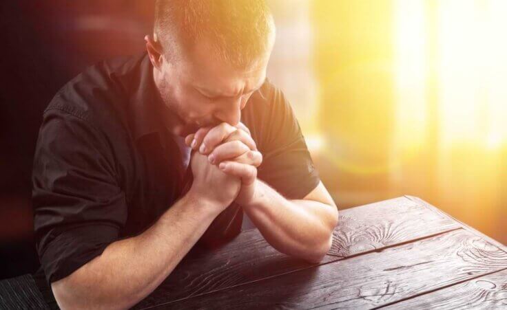 Hands clasped in front of him with his elbows on a wooden table, a man earnestly prays. © By BillionPhotos.com/stock.adobe.com