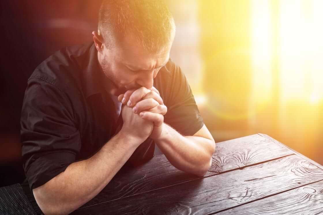 Hands clasped in front of him with his elbows on a wooden table, a man earnestly prays. © By BillionPhotos.com/stock.adobe.com