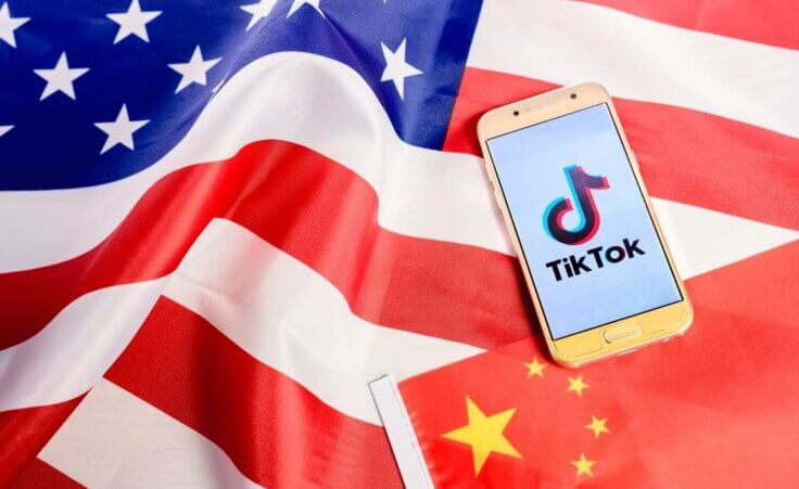 A smartphone displaying the TiTok logo sits atop a Chinese flag and an American flag, illustrative of the long-discussed TikTok ban that may occur in the US. © By Joaquin Corbalan/stock.adobe.com